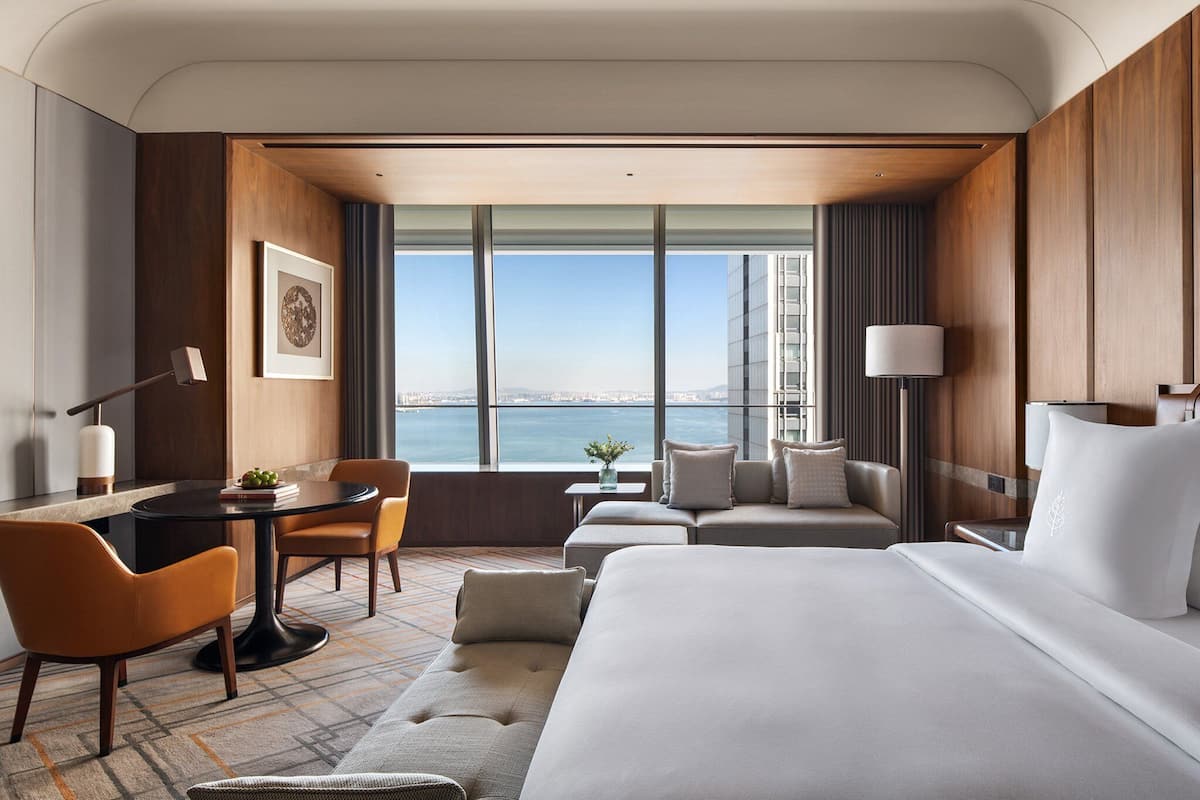 Habitación de hotel con una cama grande, un sofá, una mesa de comedor con sillas, una mesita con un florero y vistas a la ciudad y al agua.