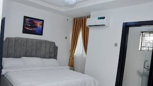 A bedroom with a bed, a nightstand, an air conditioning unit, a window with curtains, and a door leading to a bathroom. - HABBOT LUXURY APARTMENT  (Ibadan)