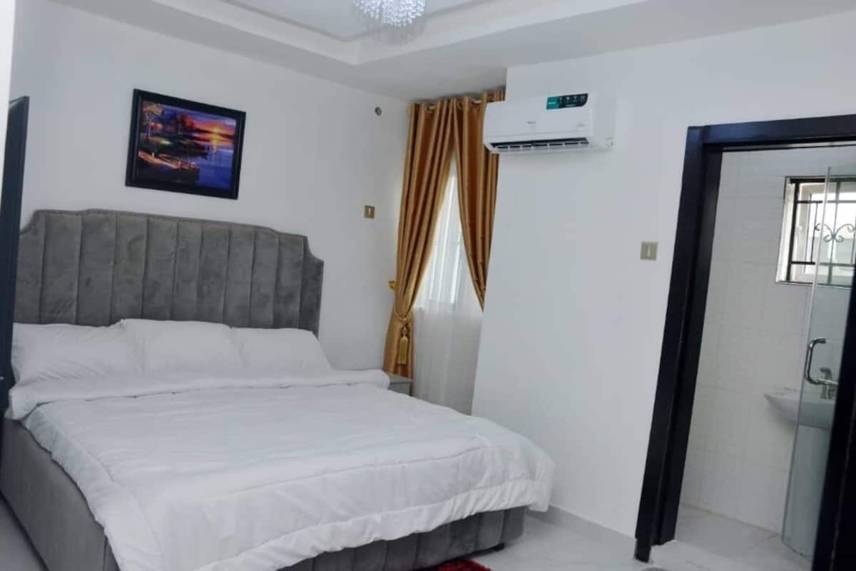 A bedroom with a bed, a nightstand, an air conditioning unit, a window with curtains, and a door leading to a bathroom.