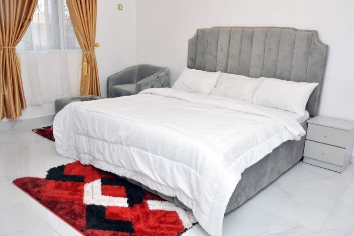 A modern bedroom with a grey upholstered bed, white bedding, a red and black rug, a grey chair, and a white nightstand.