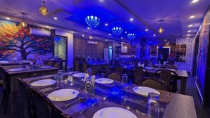 A restaurant with multiple dining tables set for guests, featuring a modern chandelier and a colorful mural on the wall. - The Pelican Beach Resort Mandarmoni (Kanthi)