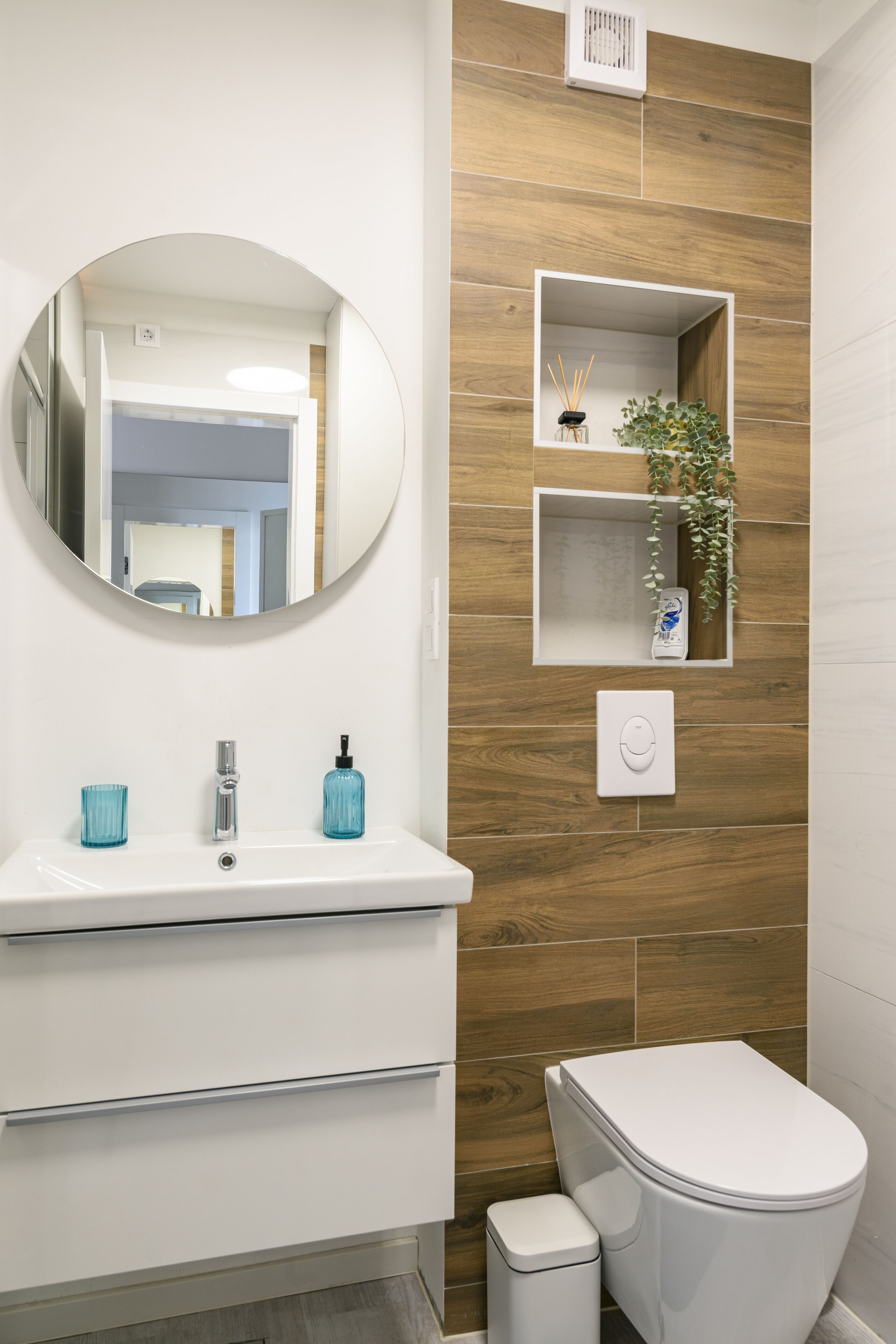A modern bathroom with a white sink, mirror, and toilet. There are wooden wall panels and a small shelf with a plant and a bottle.