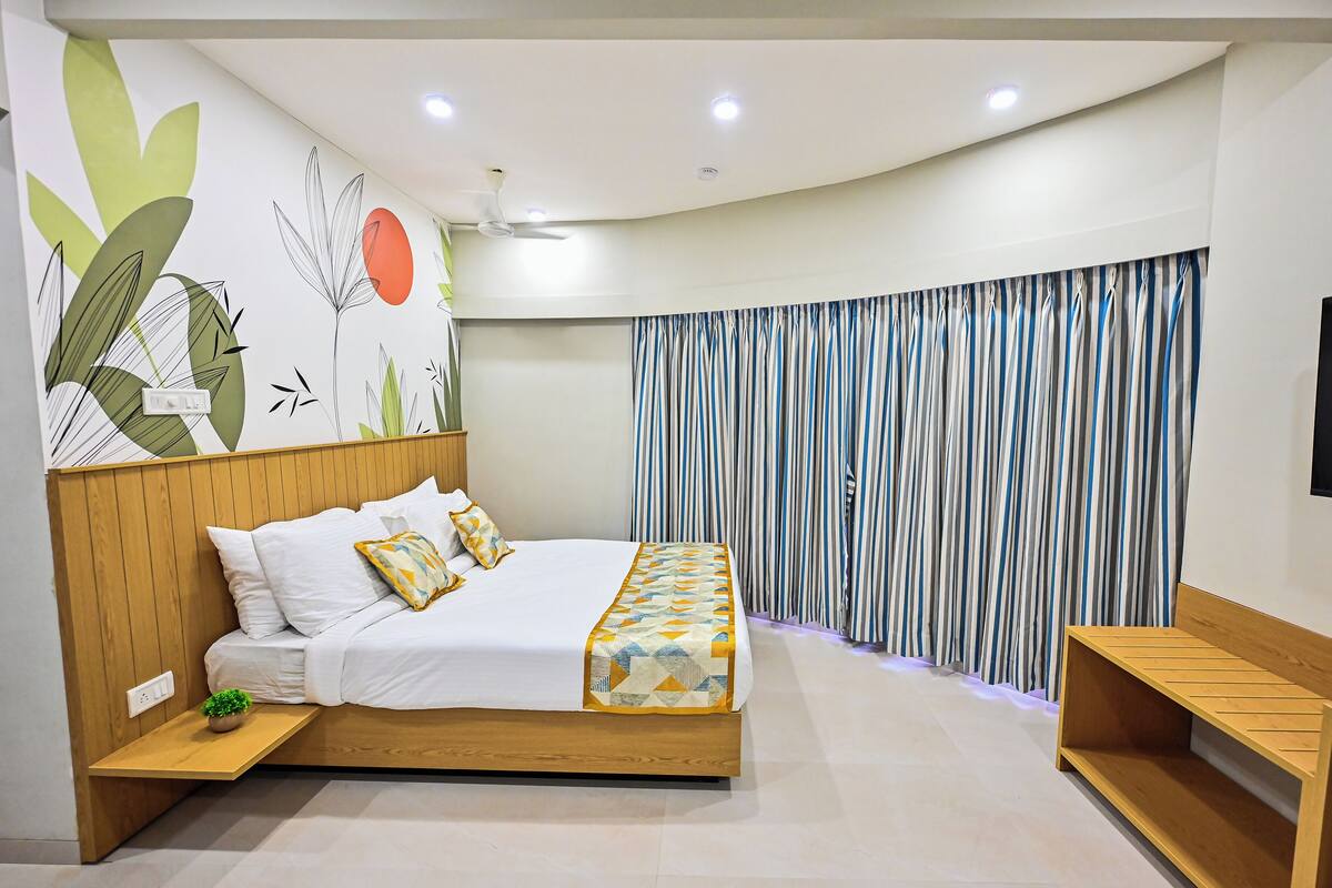 A modern bedroom with a wooden headboard, a bed with white bedding and patterned pillows, a bedside table with a plant, and a large window with striped curtains.