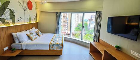 A modern hotel room with a large bed, a TV, a wooden bench, and a view of the city.