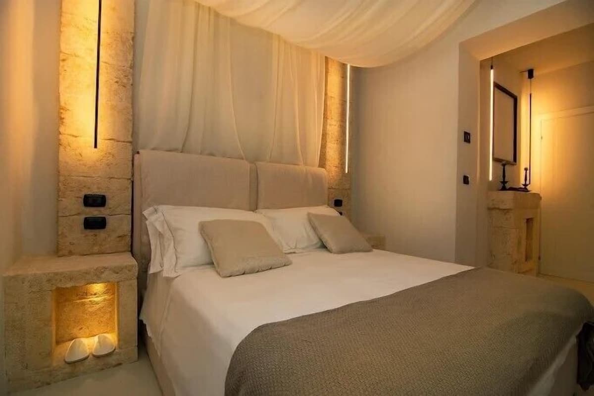 A bedroom with a stone wall, a bed with white linens, a bedside table with a lamp, and a mirror on the wall.