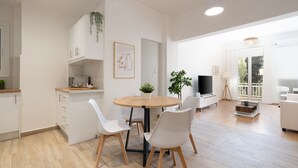 A modern kitchen with white cabinets, a wooden dining table, and white chairs. There is a potted plant on the counter and a framed picture on the wall.