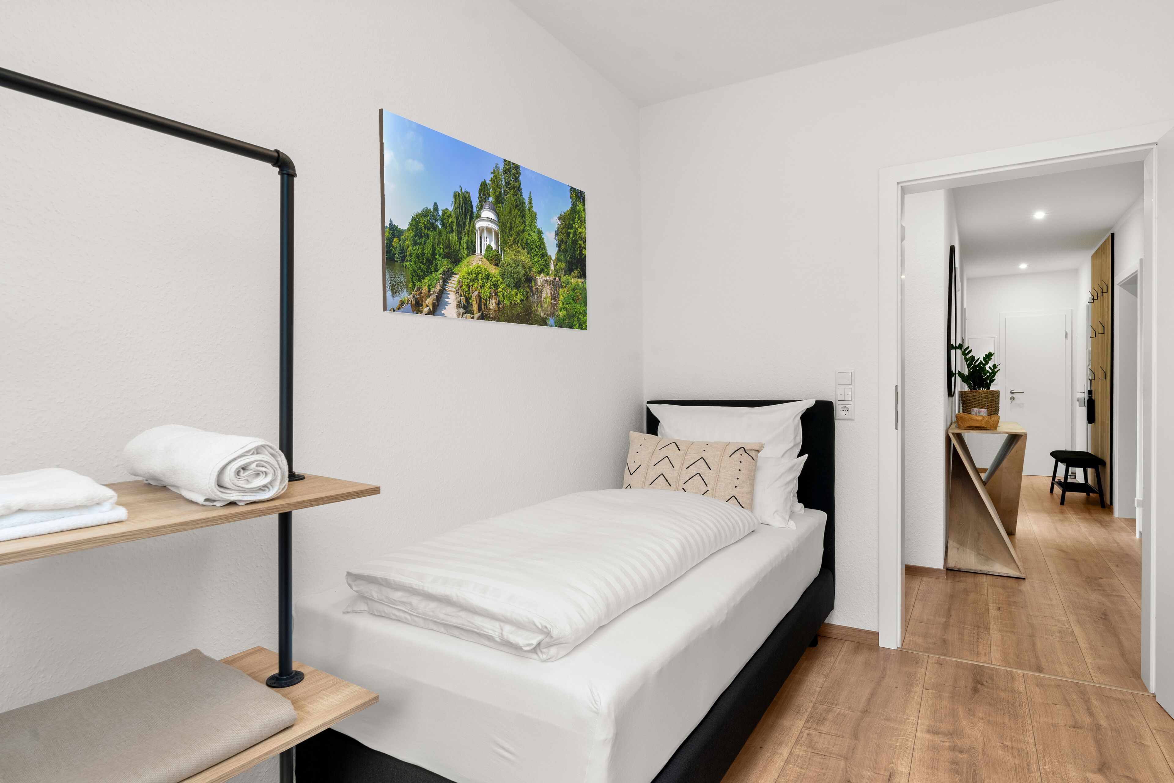 Une chambre à coucher moderne avec un lit, une étagère avec des serviettes, un tableau au mur et une vue sur une autre pièce.