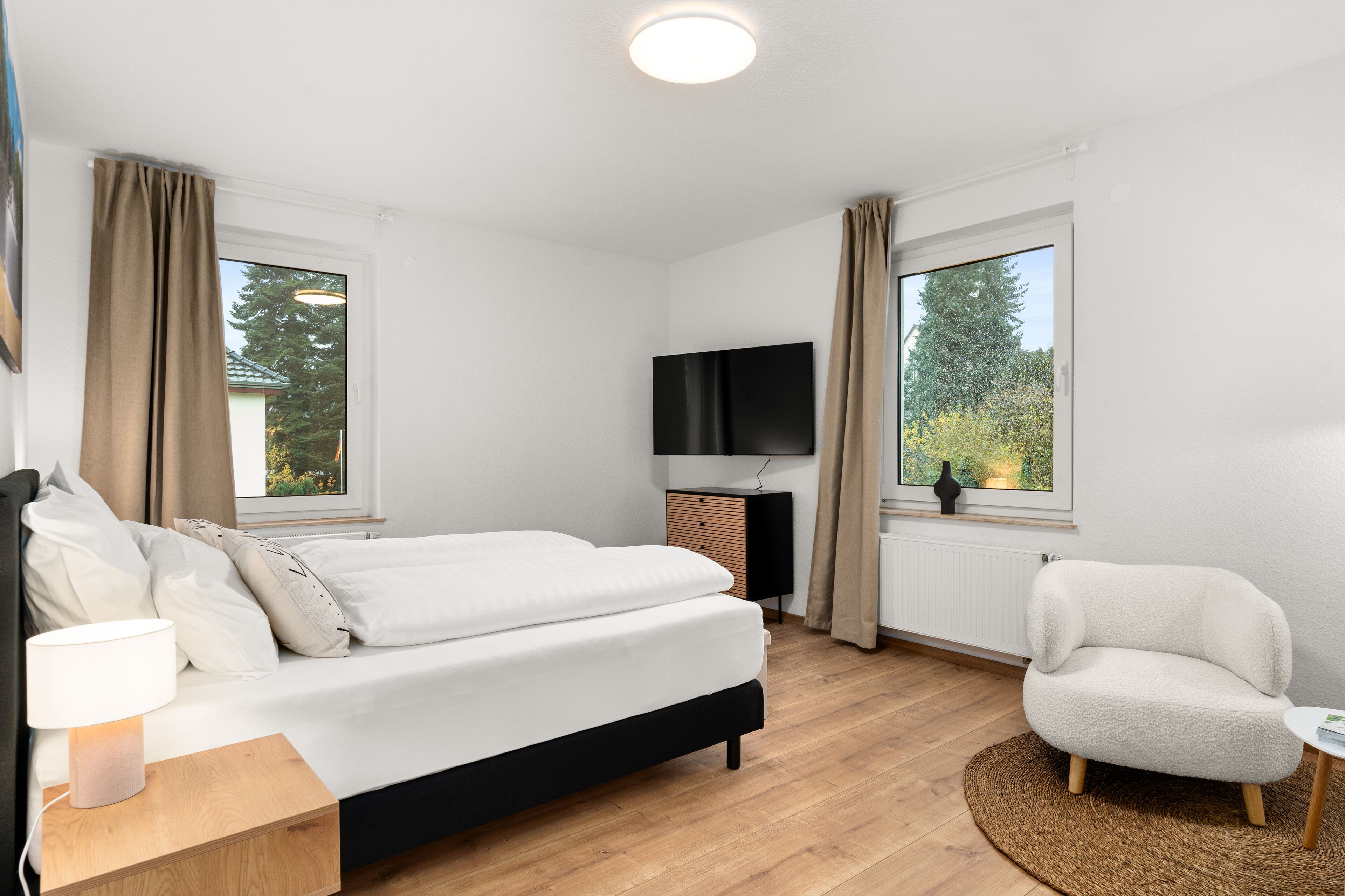 Ein modernes Schlafzimmer mit Bett, Nachttisch, Stuhl, Fernseher und Fenstern.