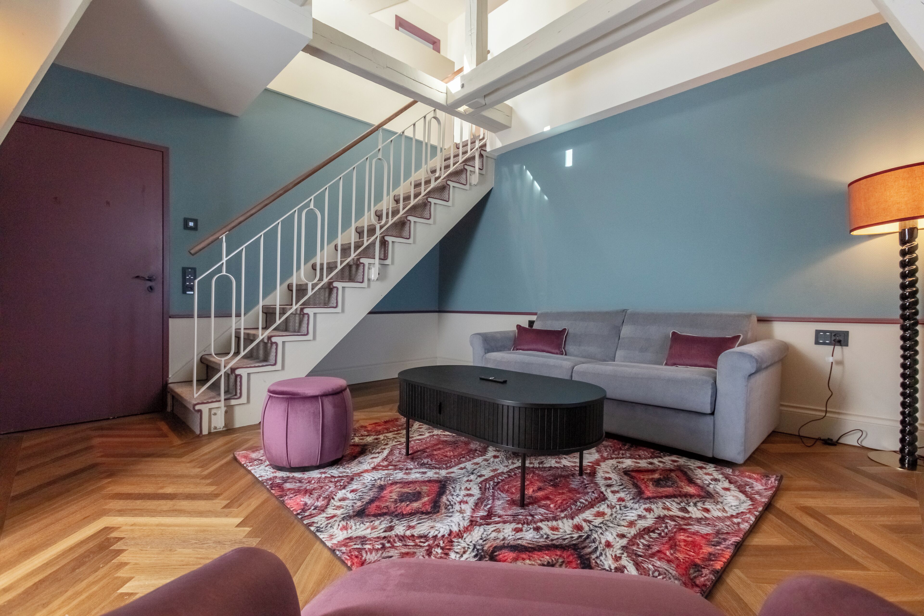 A modern living room with a staircase, a grey sofa, a patterned rug, and a purple ottoman.