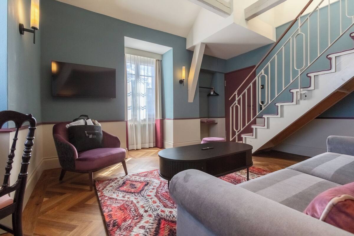 Un salon moderne avec un canapé, un fauteuil et une table basse. On y trouve un escalier avec une rampe et une porte qui mène à une autre pièce.