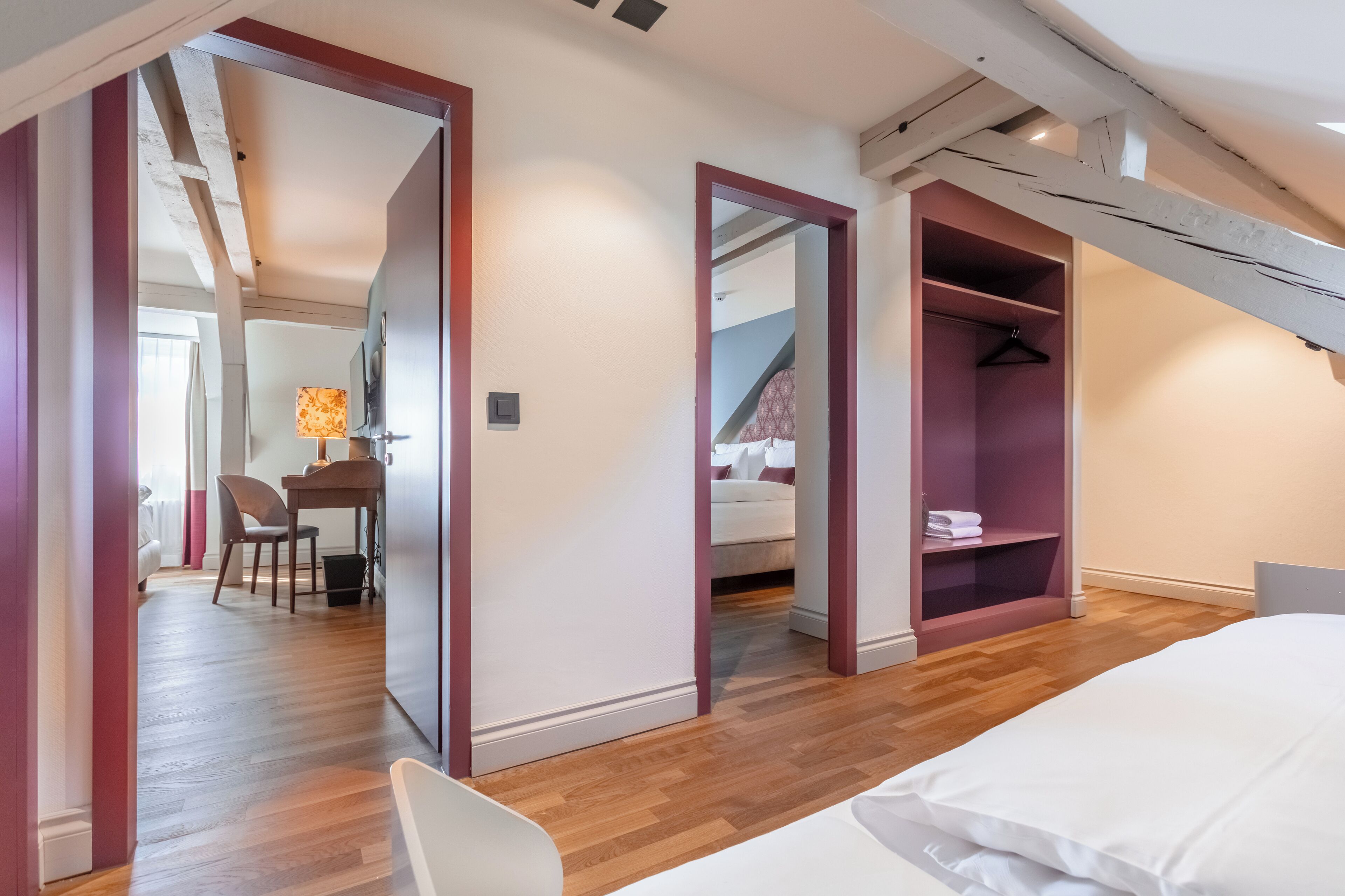 A modern bedroom with a wooden floor, a bed with white bedding, a desk with a lamp, and a wardrobe with hanging space.