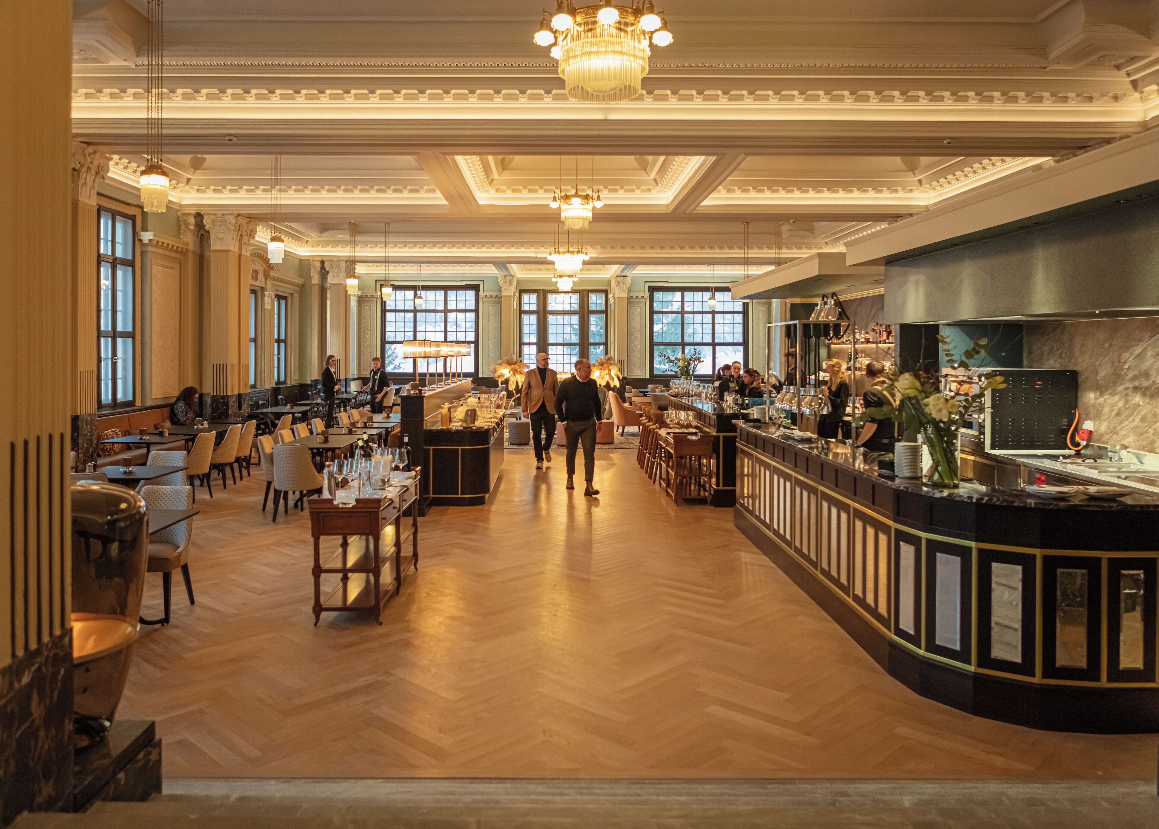 A spacious hotel lobby with a bar area, wooden flooring, and large windows.