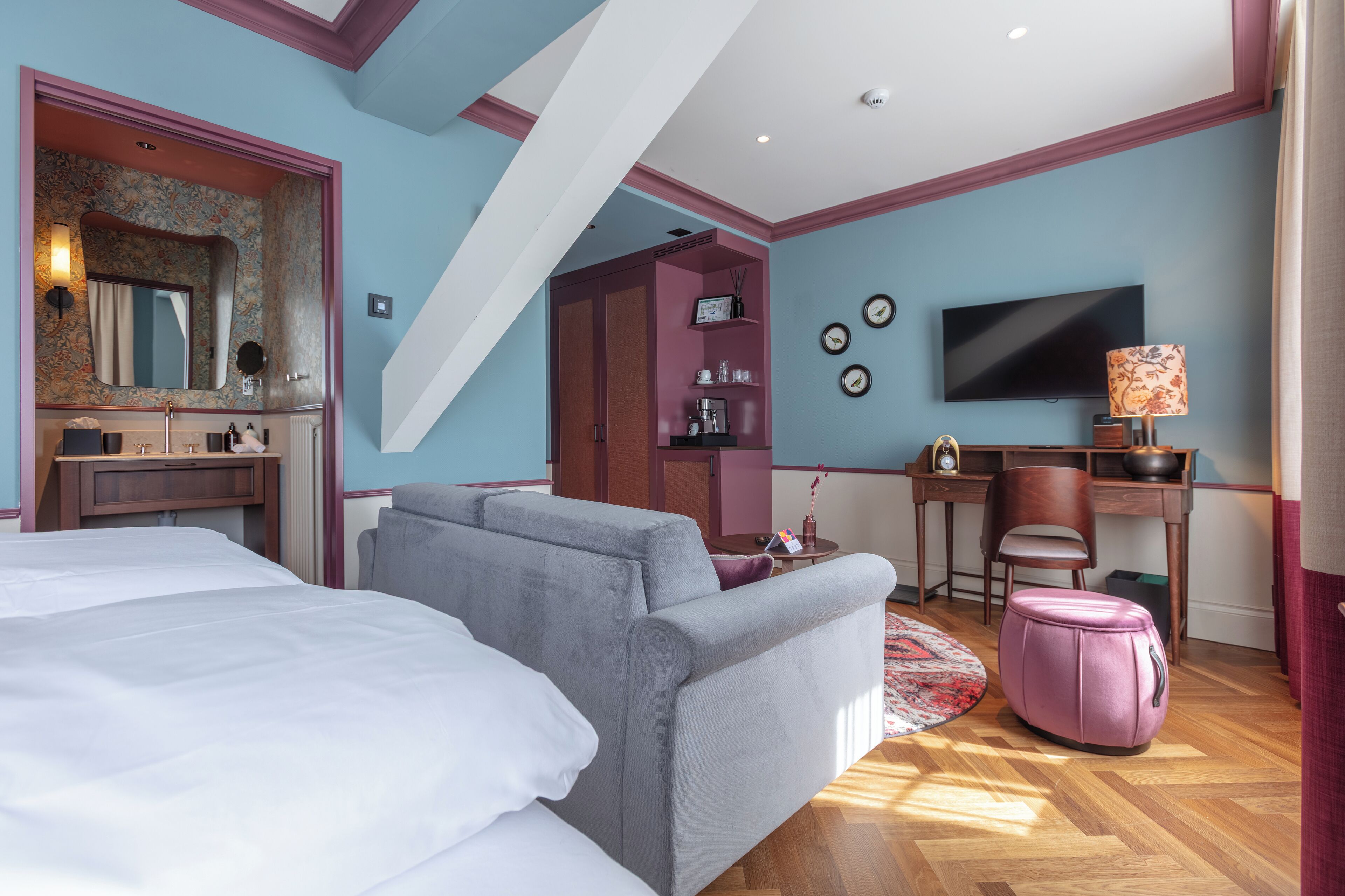 A hotel room with a bed, a sofa, a desk with a chair, a television, and a small kitchenette area.