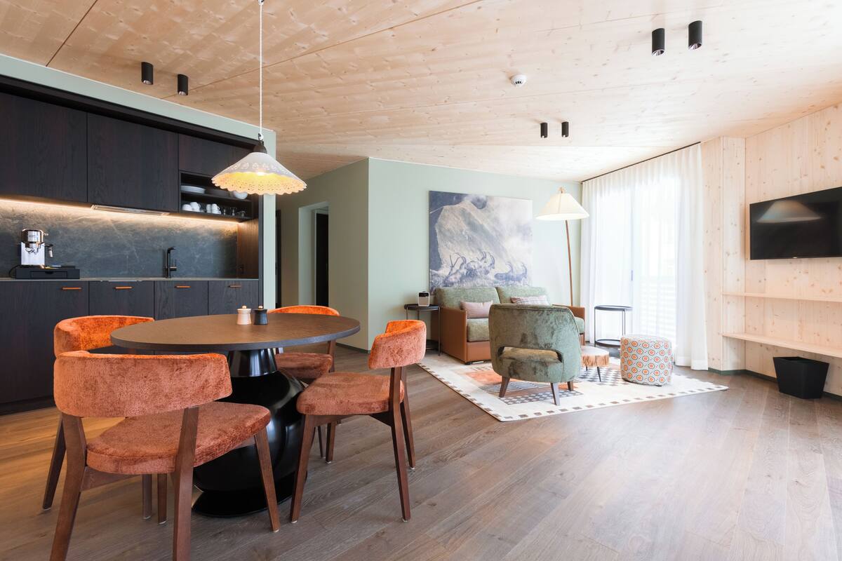 Une cuisine moderne avec des meubles de couleur sombre, une table de salle à manger ronde entourée de chaises orange, un canapé et un téléviseur fixé au mur.