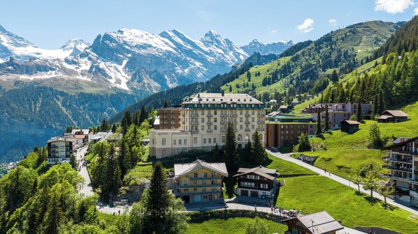 Een bergresort met een groot hotel, weelderige groene omgeving en besneeuwde bergtoppen op de achtergrond.