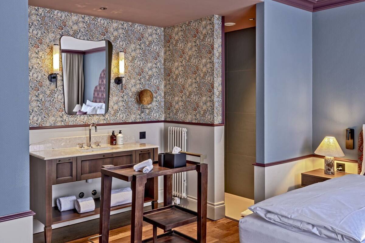 Une chambre d’hôtel avec un plancher en bois, un lit, un bureau avec une chaise, une coiffeuse avec un miroir et une salle de bain avec une douche.