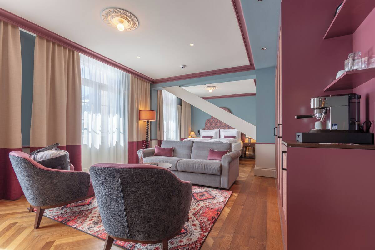 Un salon moderne avec un canapé gris, deux fauteuils, une machine à café et un escalier menant à un autre niveau.