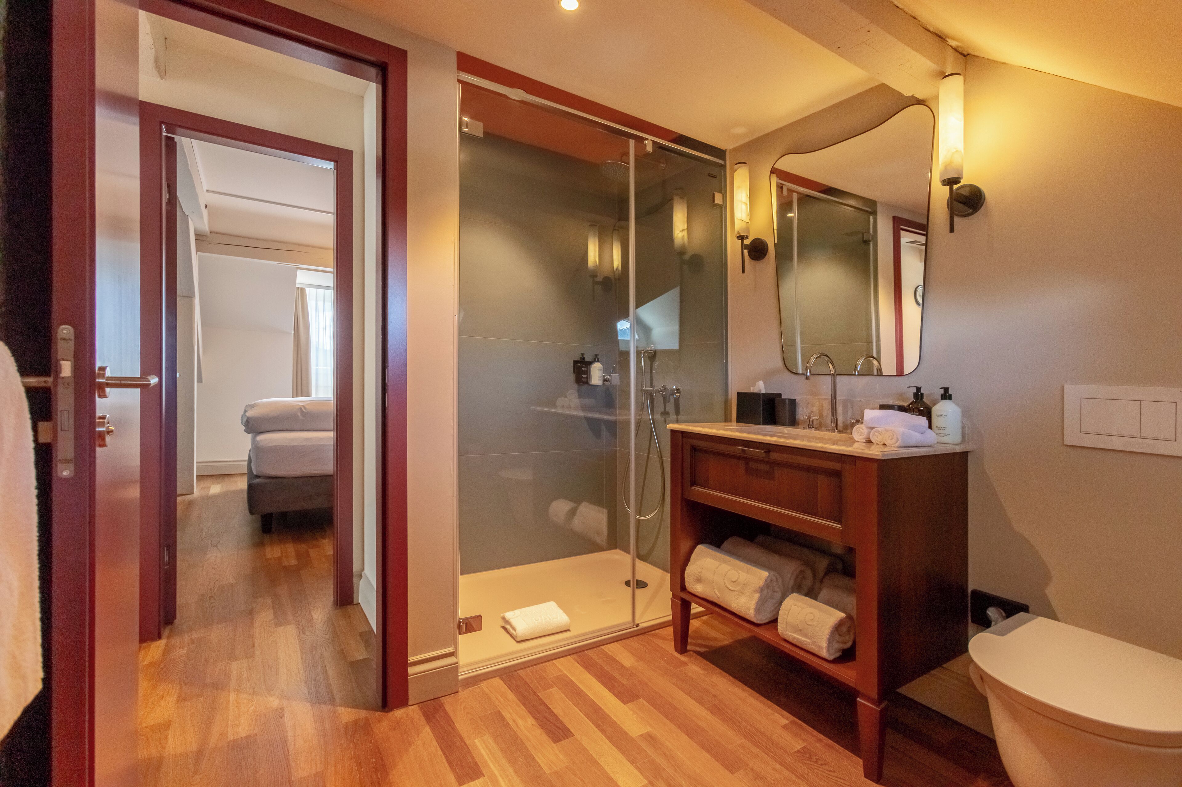 Un baño moderno con ducha de vidrio, tocador de madera y una habitación con cama a la vista.