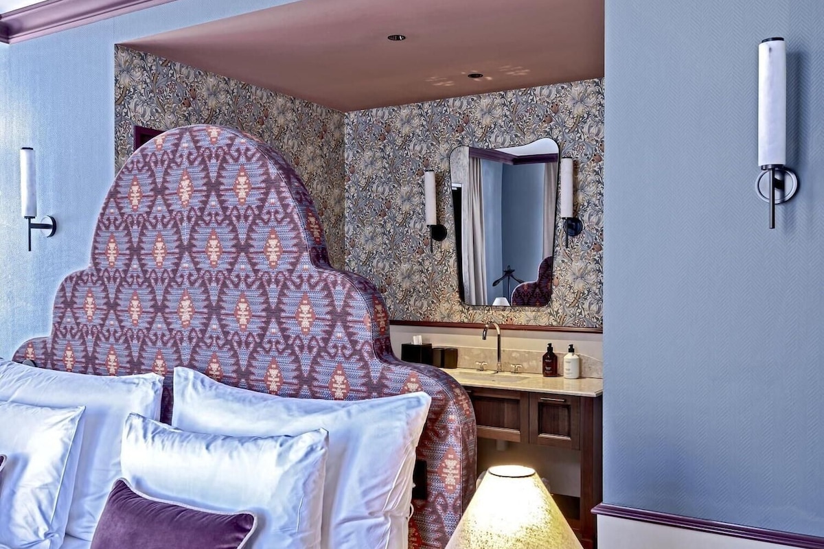 Quarto com cabeceira estampada, mesa de cabeceira com abajur, espelho e toucador com artigos de higiene pessoal.