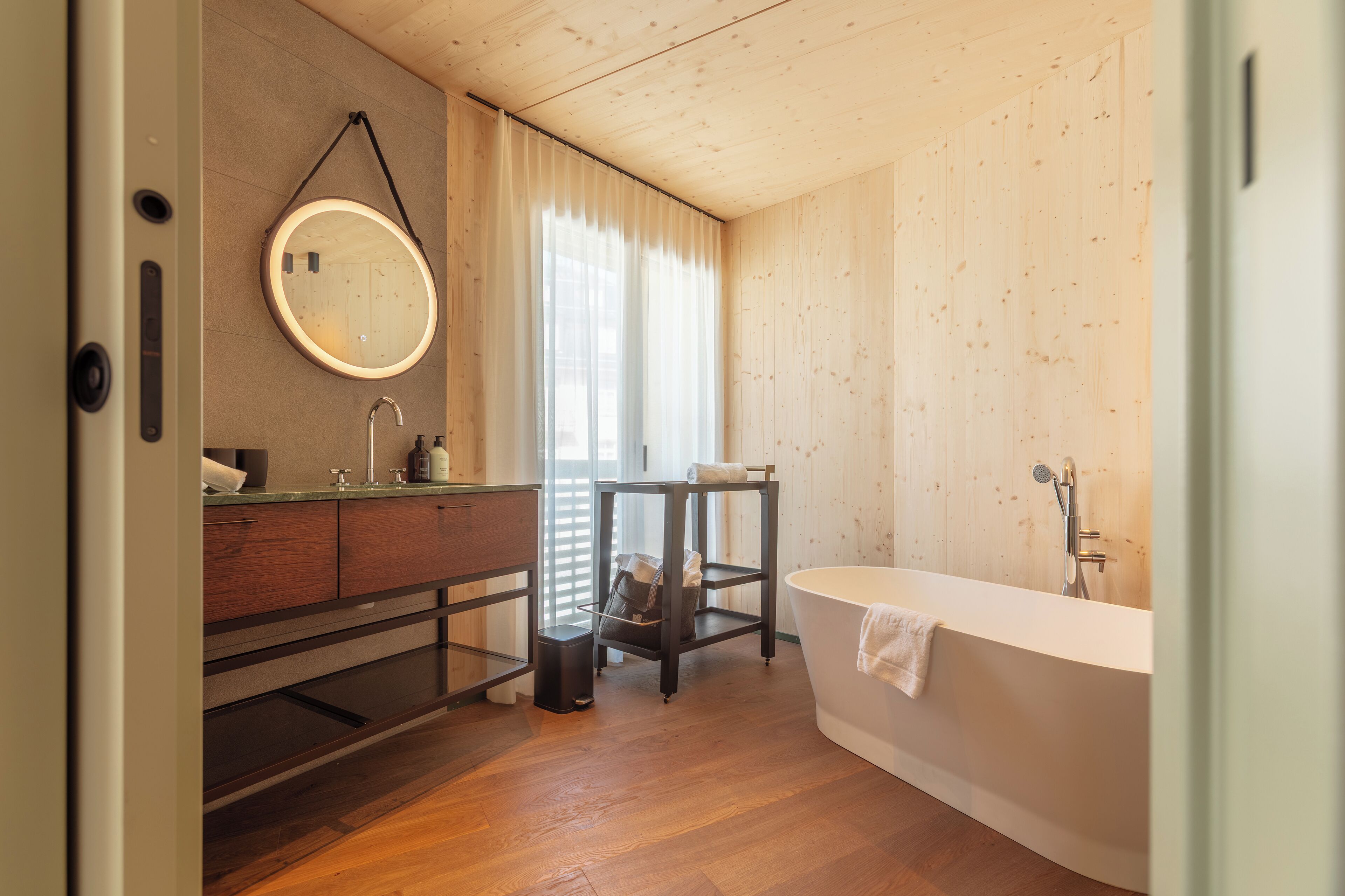 A modern bathroom with a wooden floor, a round mirror, a white bathtub, and a wooden vanity.
