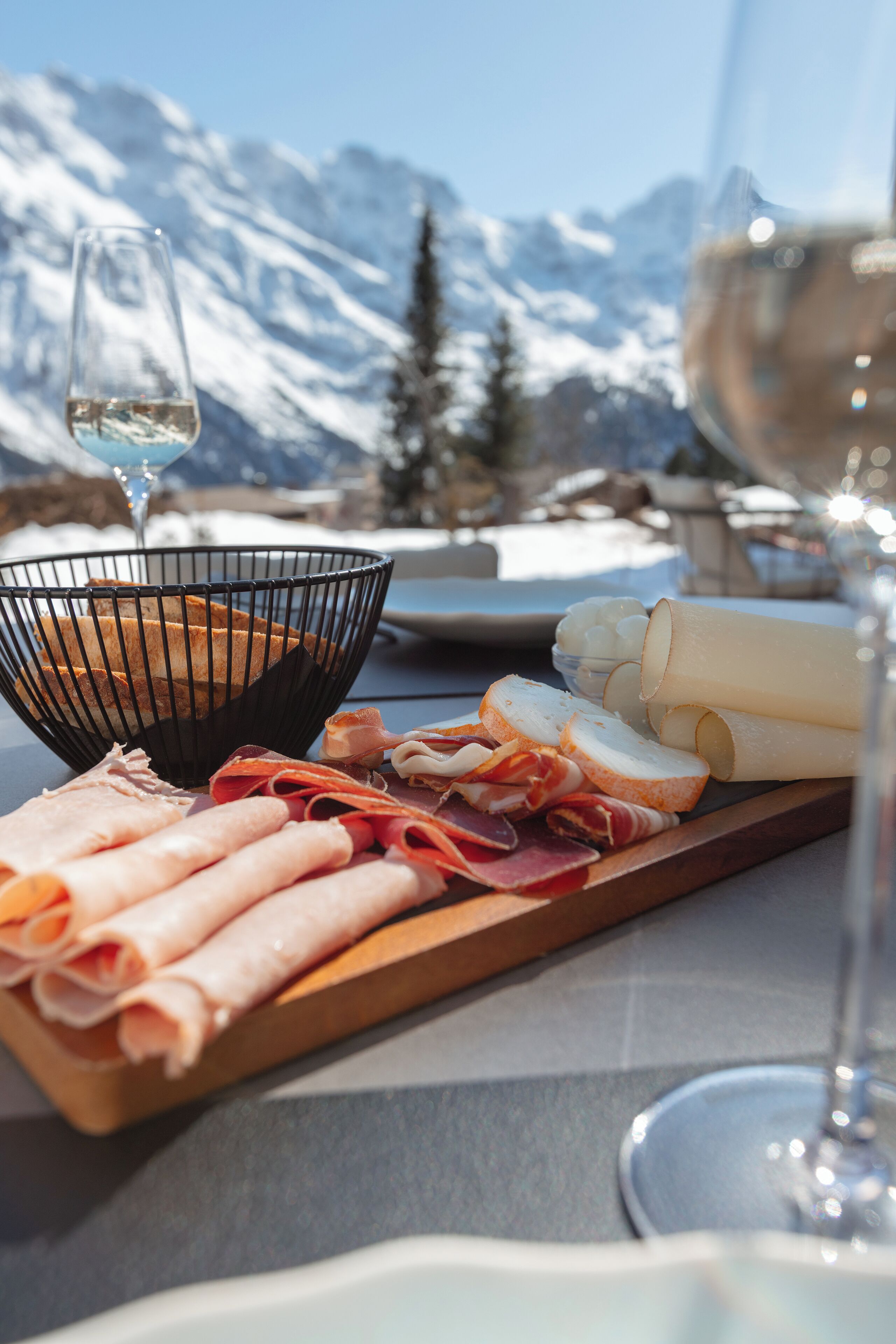 Una tabla de fiambres con diferentes carnes y quesos, una copa de vino blanco y una vista de montañas nevadas al fondo.