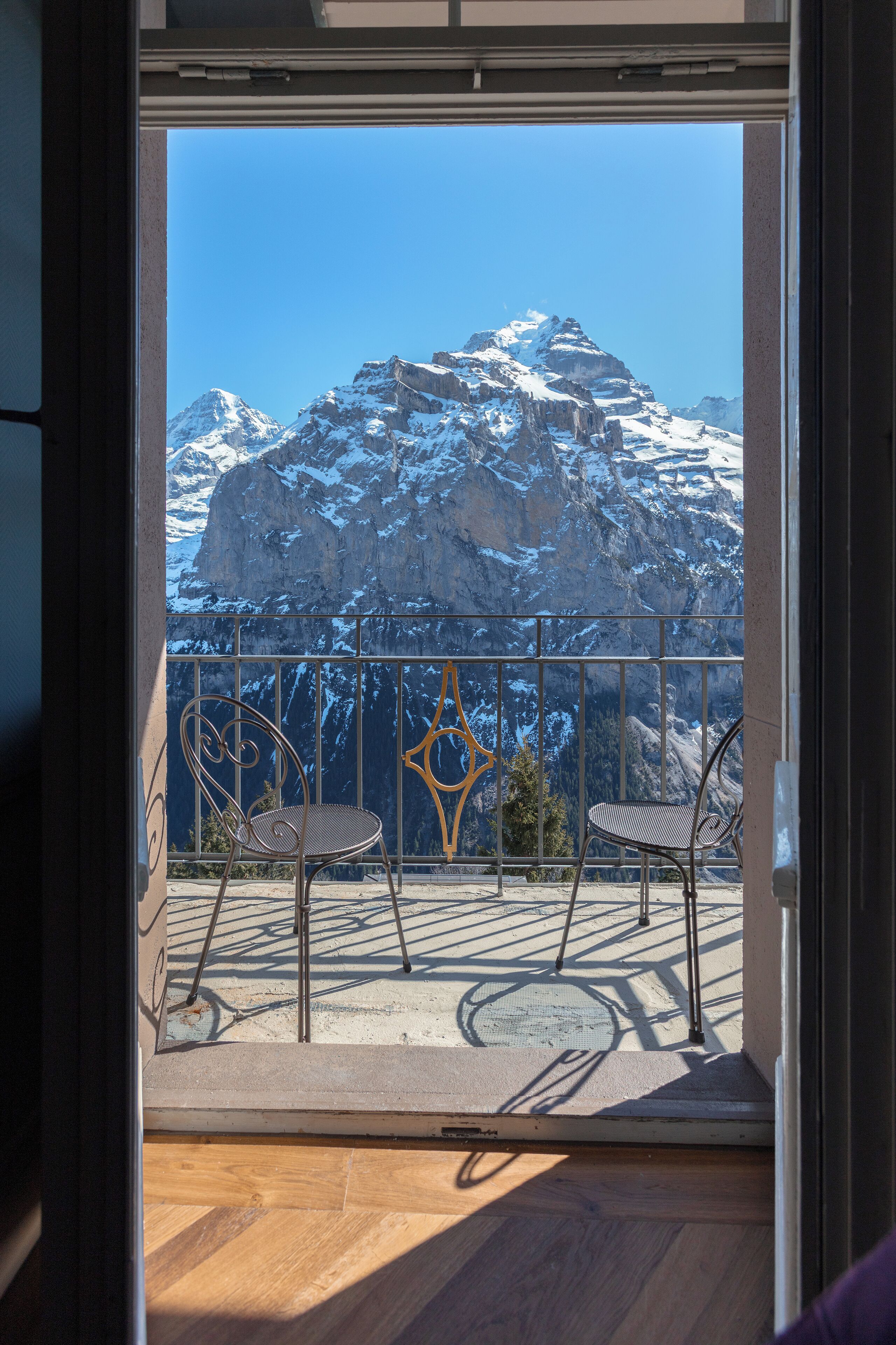 A balcony with two chairs and a metal railing, overlooking a snow-covered mountain range.
