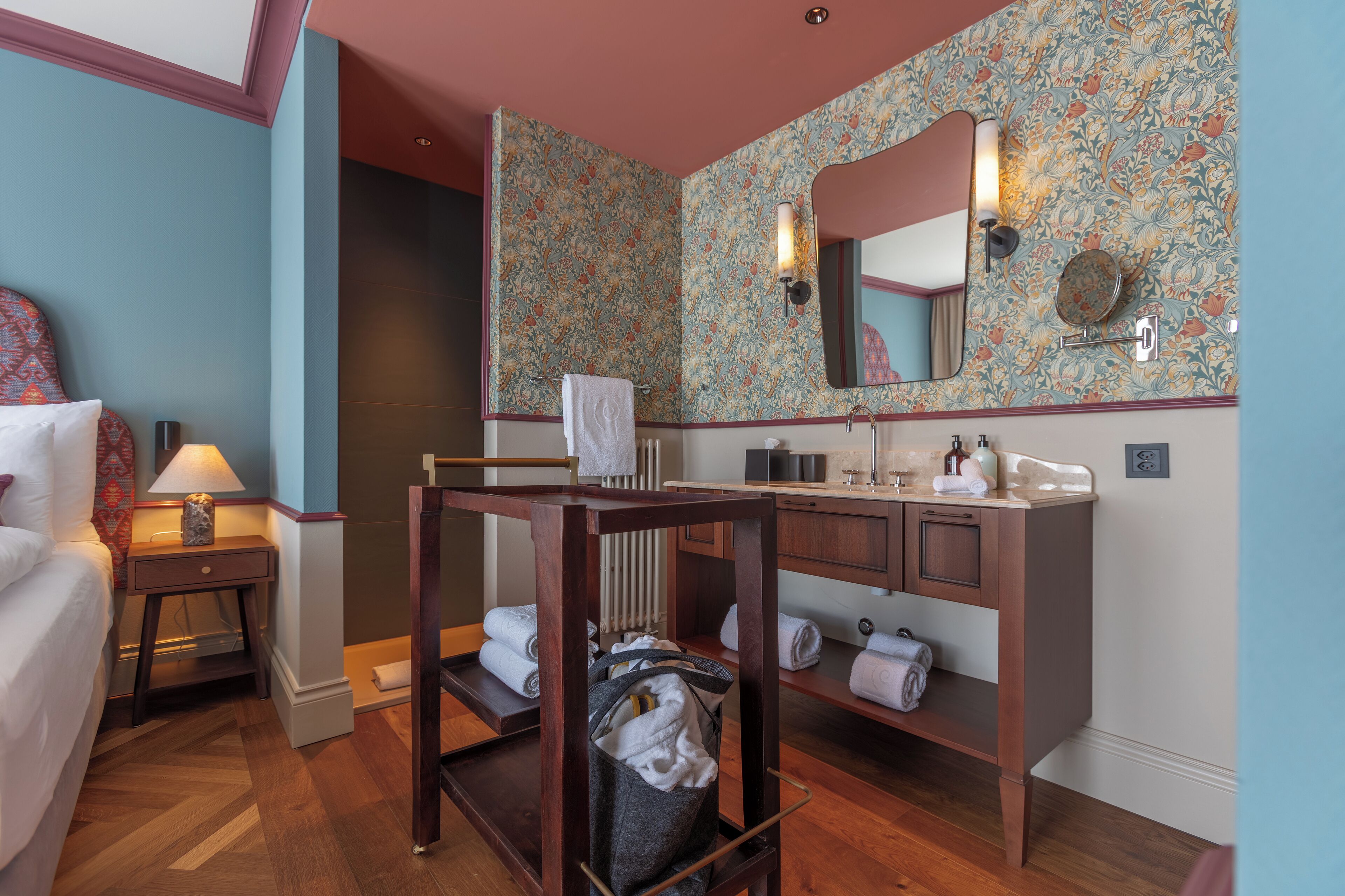 Ein Hotelzimmer mit einer Holzbank, einem kleinen Tisch mit Lampe, einem großen Spiegel und einem Badezimmer-Waschtisch mit Handtüchern.