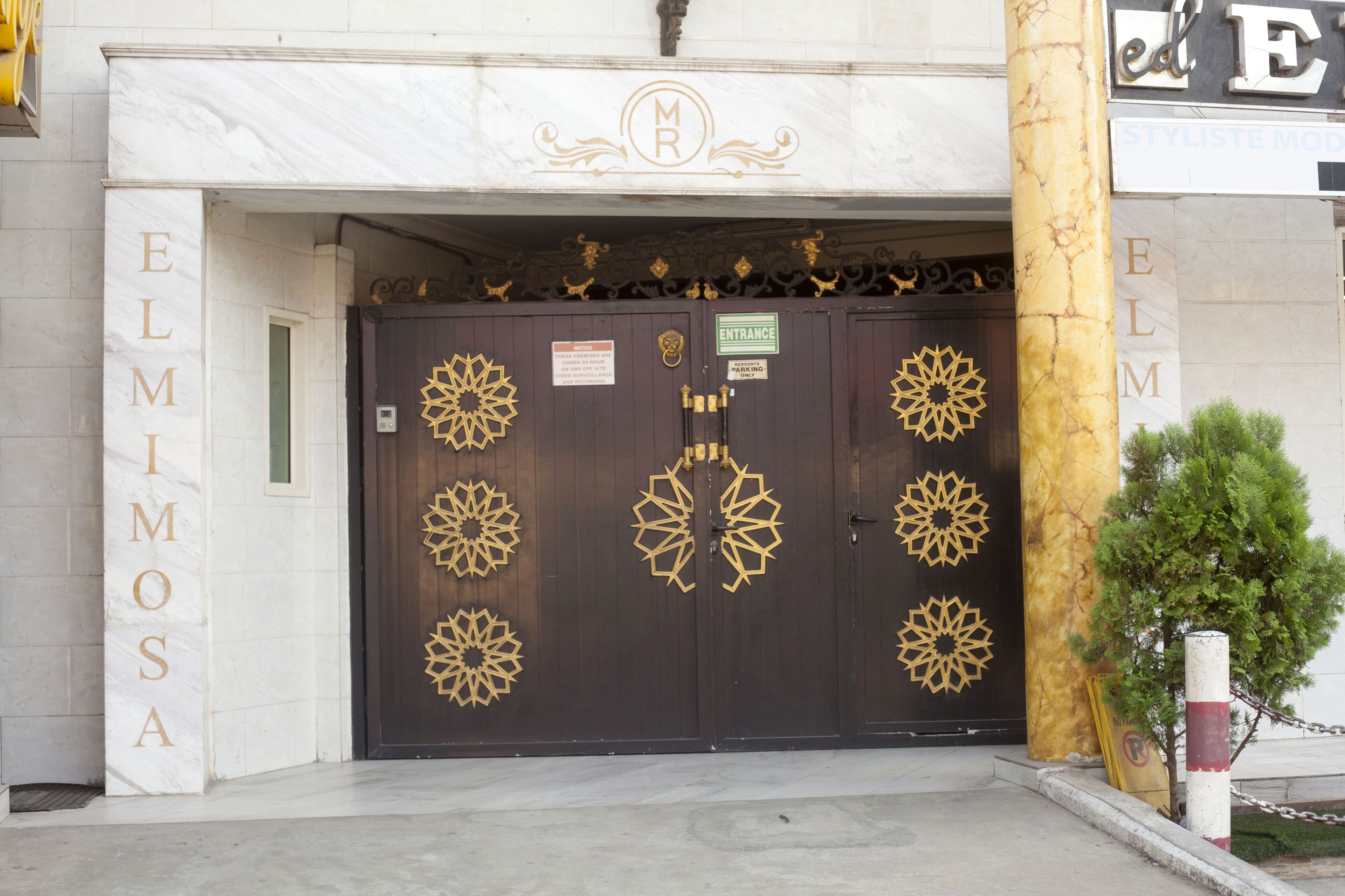 Une entrée d’immeuble avec une porte marron foncé ornée de motifs géométriques dorés et le nom « ELMOSA » inscrit sur le mur.