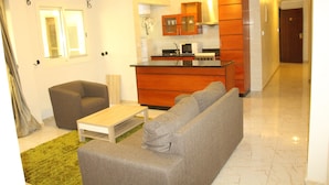 A modern living room with a grey sofa, a small coffee table, and a brown armchair. A kitchen with wooden cabinets and a microwave is visible in the background.