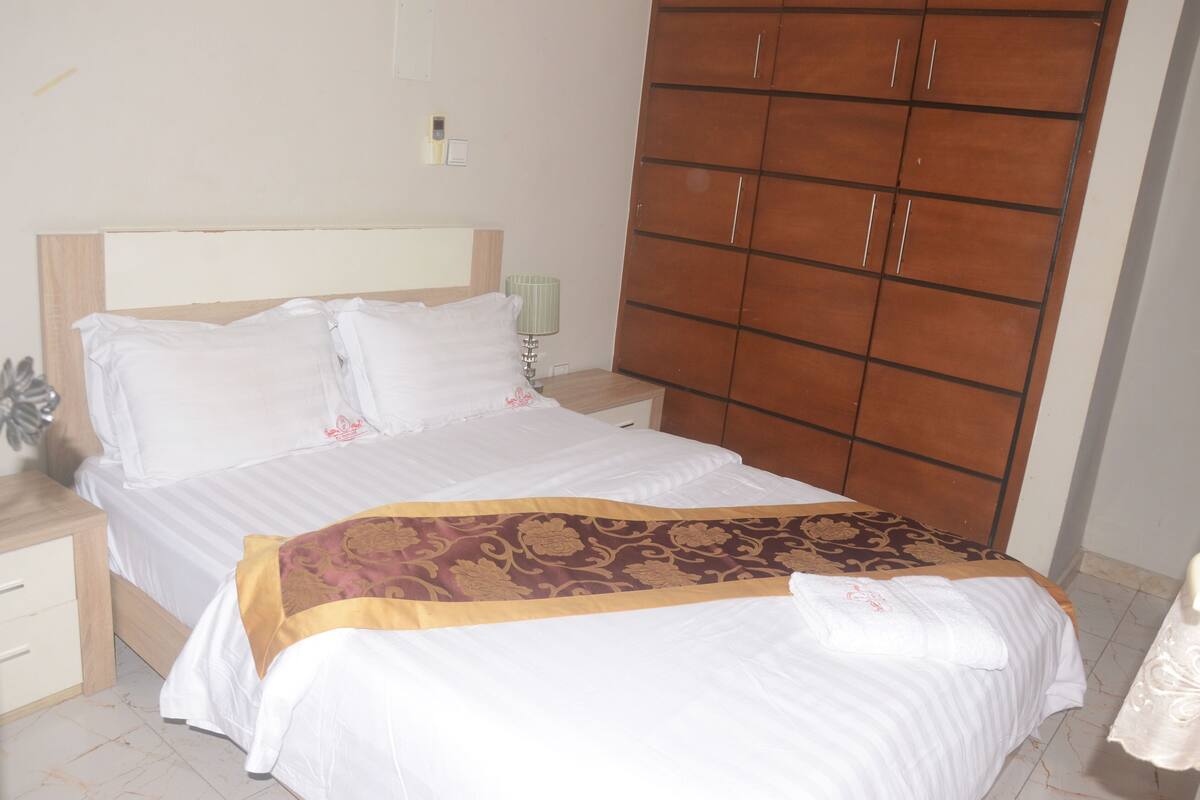 A neatly made bed with a patterned bedspread, a wooden wardrobe, and a bedside table with a lamp.