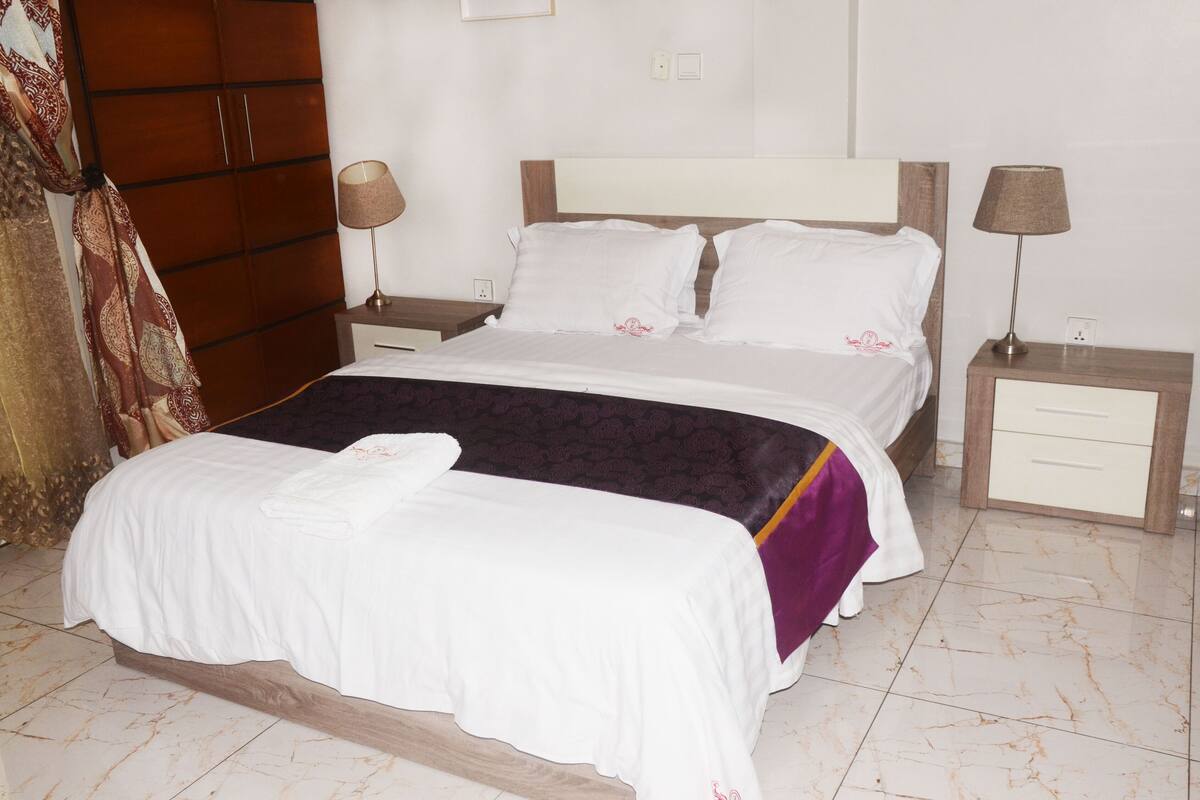 A double bed with white bedding, a wooden headboard, two bedside tables with lamps, and a patterned throw blanket.