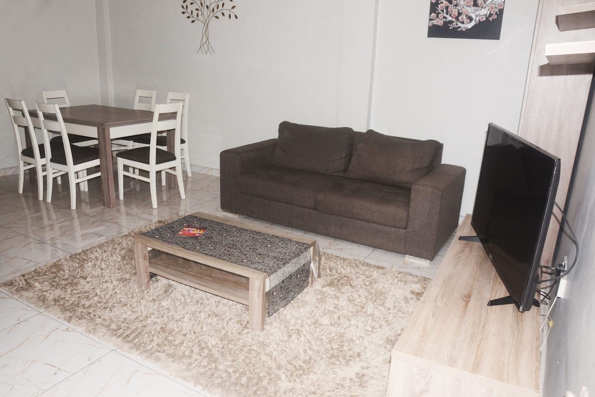 A living room with a brown sofa, a wooden coffee table, a dining table with white chairs, and a flat-screen TV.