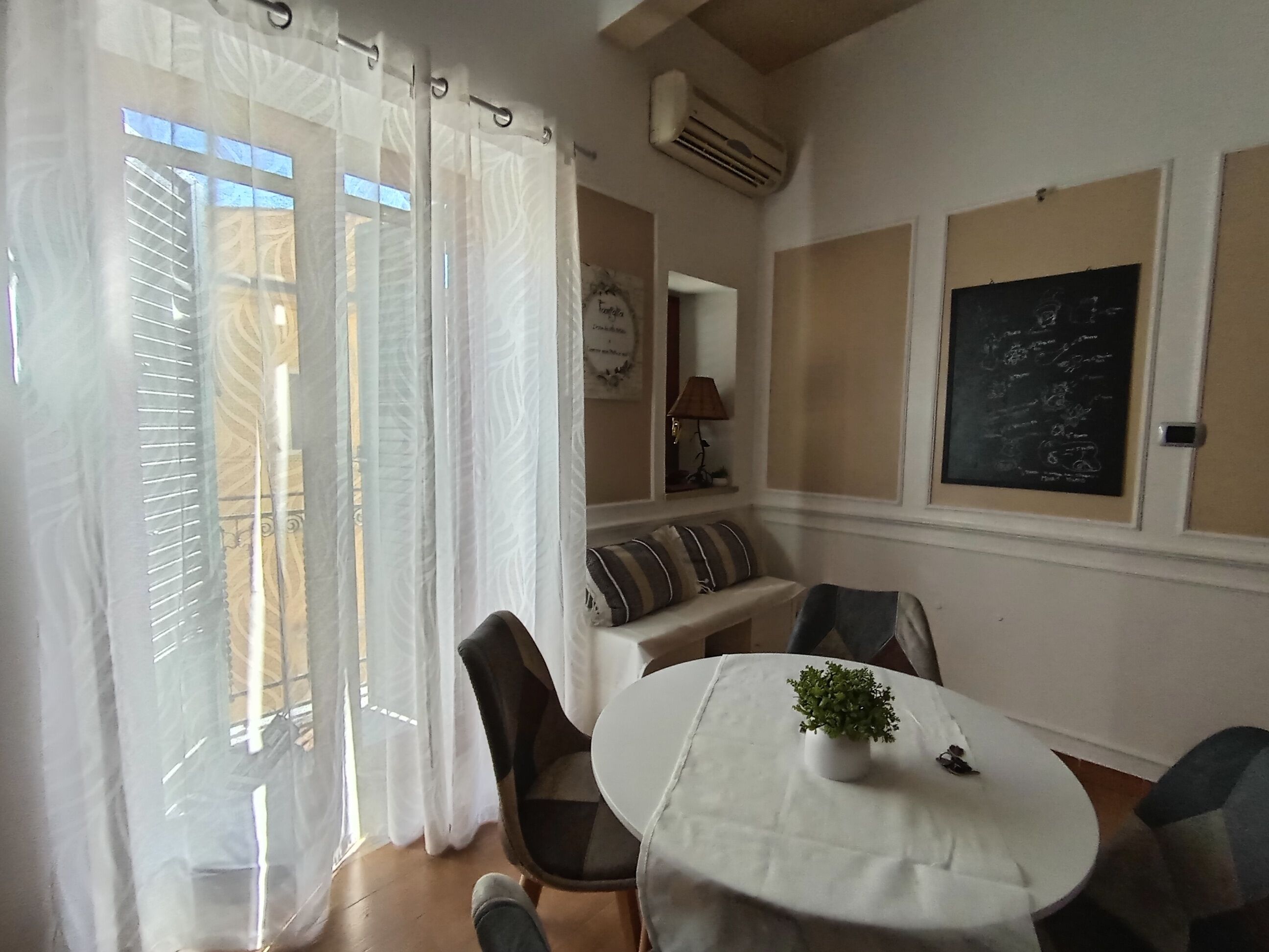 A dining area with a round table, chairs, a potted plant, and a view through a window with curtains.