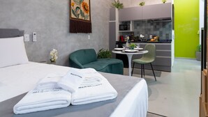 Une chambre d’hôtel comprenant un lit, un fauteuil vert, une petite table à manger et un coin cuisine.