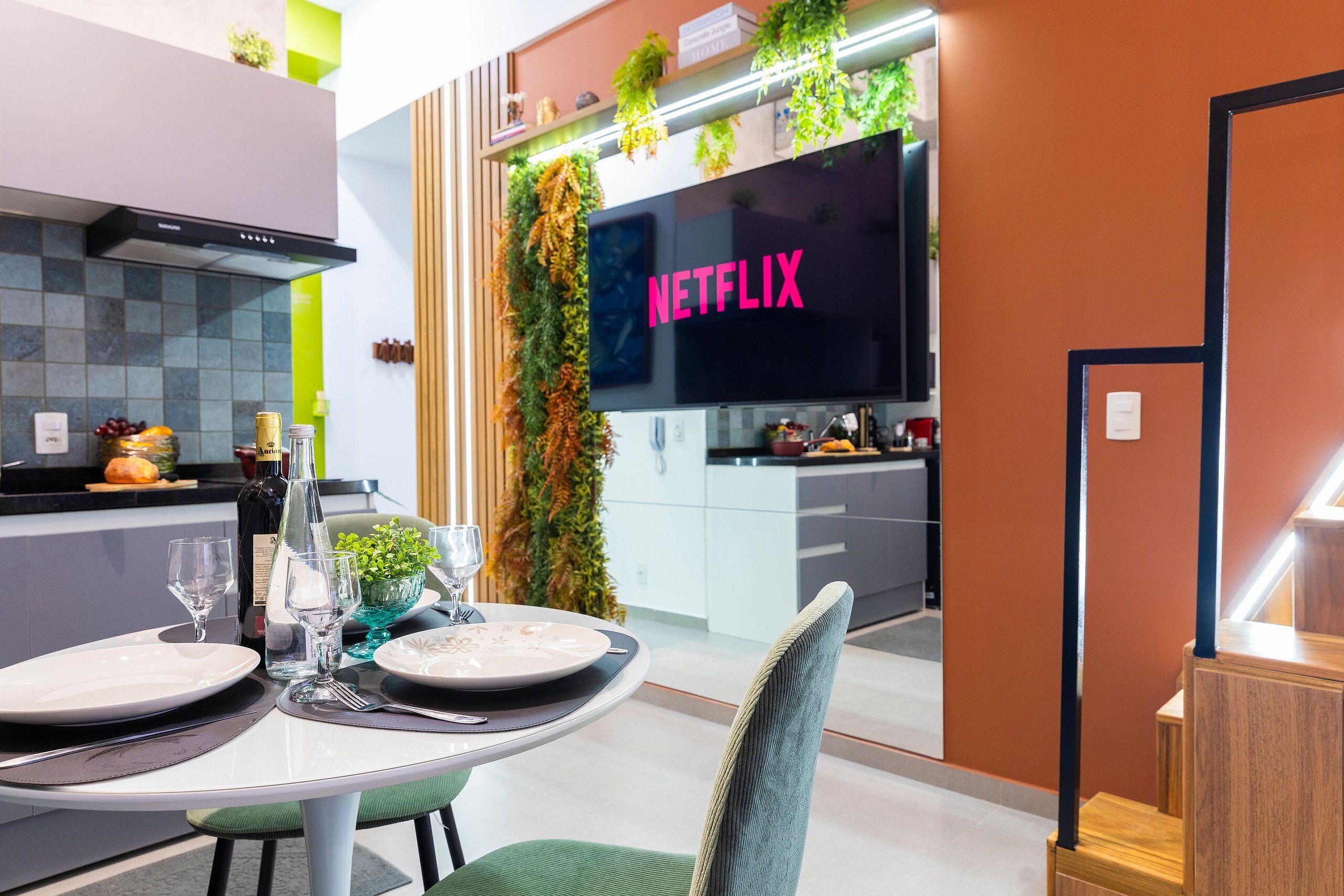 A modern kitchen with a large flat-screen TV displaying "NETFLIX" in the background, a dining area with a table set for two, and a staircase with wooden steps.