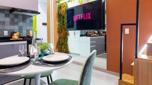 Une cuisine moderne avec un grand téléviseur à écran plat affichant le logo « NETFLIX » en arrière-plan, un coin repas avec une table dressée pour deux personnes et un escalier avec des marches en bois.