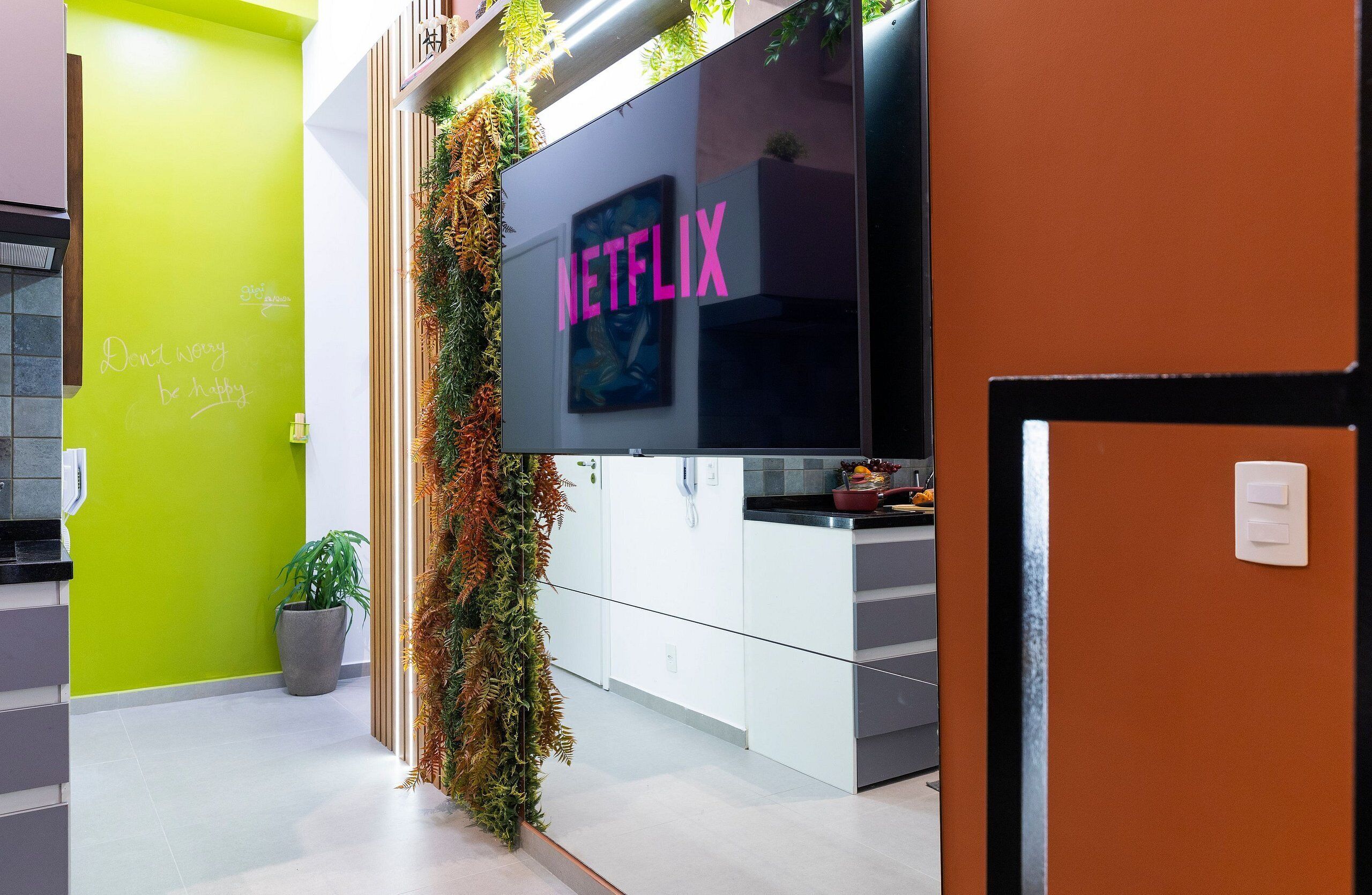 A modern kitchen with a large flat-screen TV displaying the Netflix logo, a green accent wall, and a decorative plant.