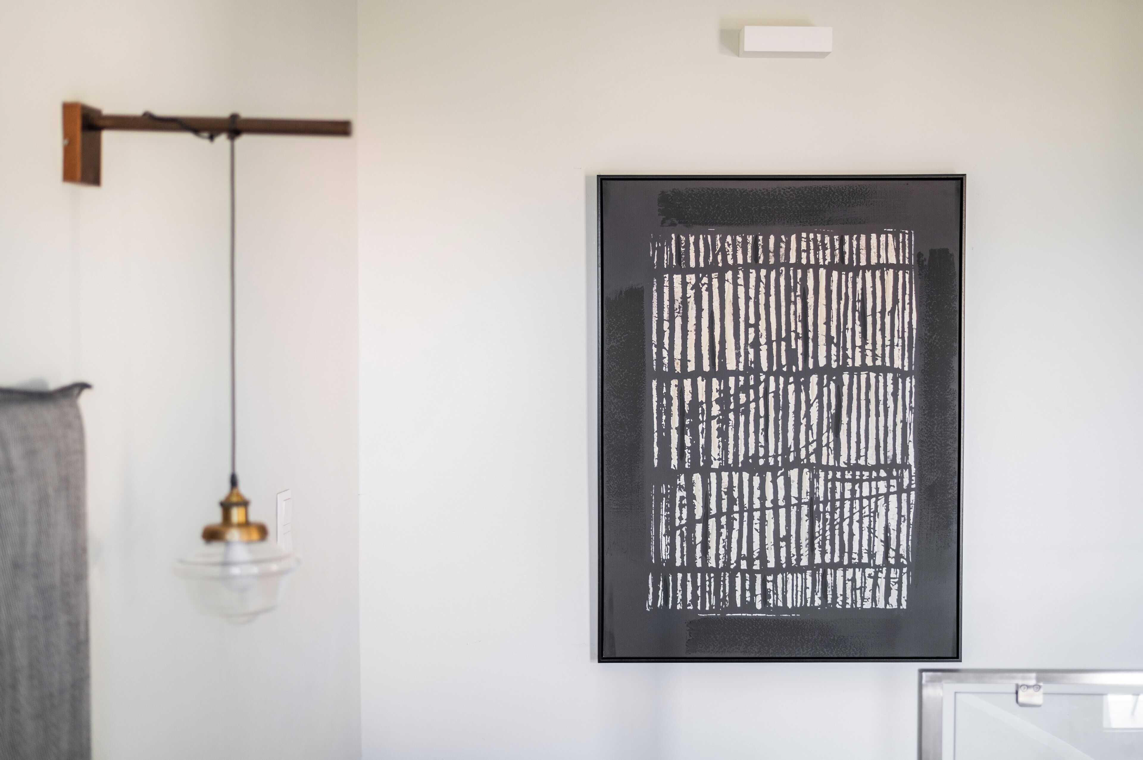 A framed abstract artwork on a white wall, a hanging pendant light, and a wooden bracket.