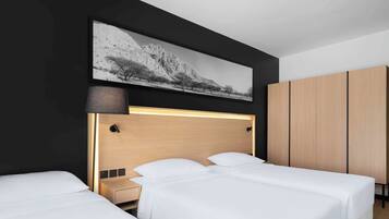 Une chambre d’hôtel avec deux lits simples, une tête de lit en bois, une table de chevet, une lampe, une armoire et une grande photographie en noir et blanc accrochée au mur.