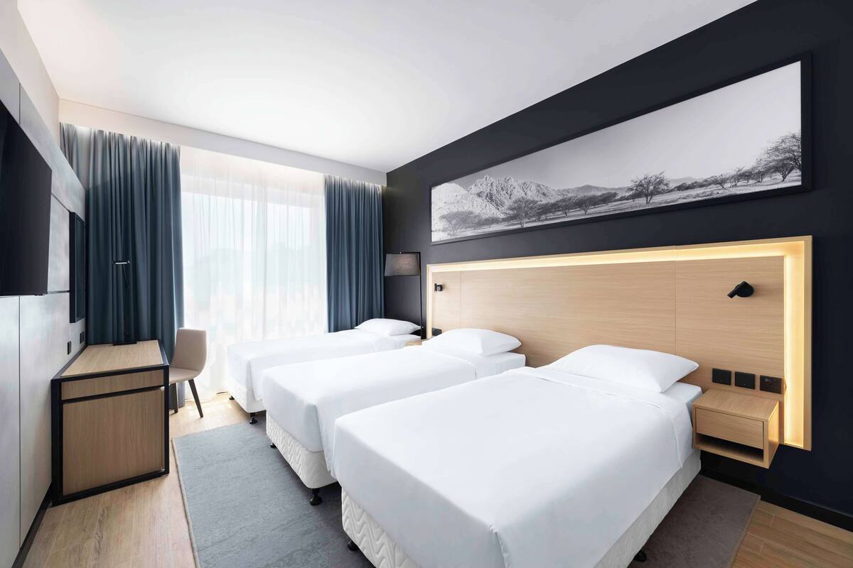 A hotel room with two single beds, a desk, a chair, a TV, and a large window with curtains.