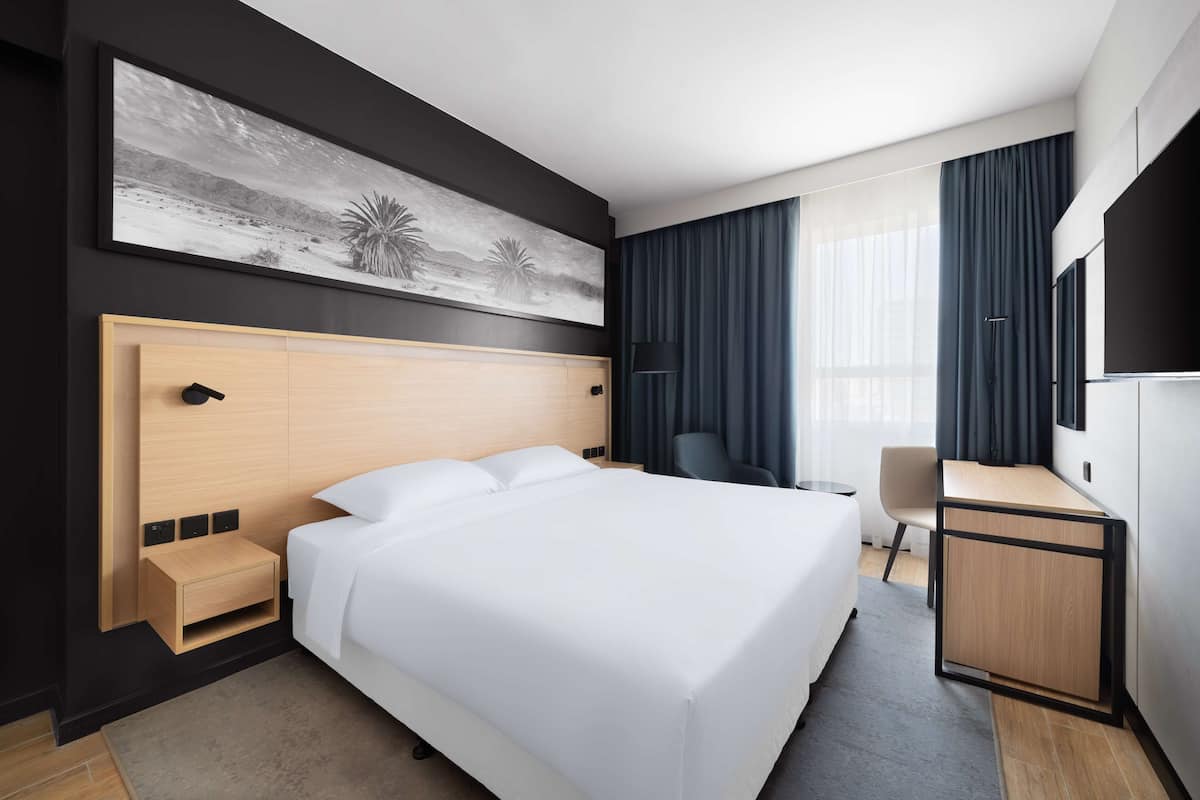 A modern hotel room with a large bed, a desk, a chair, a TV, and a painting on the wall.