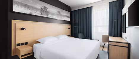 A modern hotel room with a large bed, a desk, a chair, a TV, and a painting on the wall.