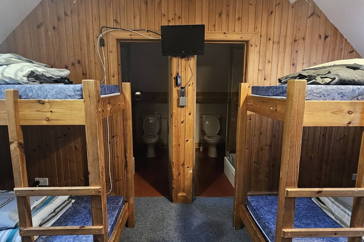 Una habitación con dos literas, una puerta de madera, un televisor instalado en la pared y un baño visible a través de la puerta.