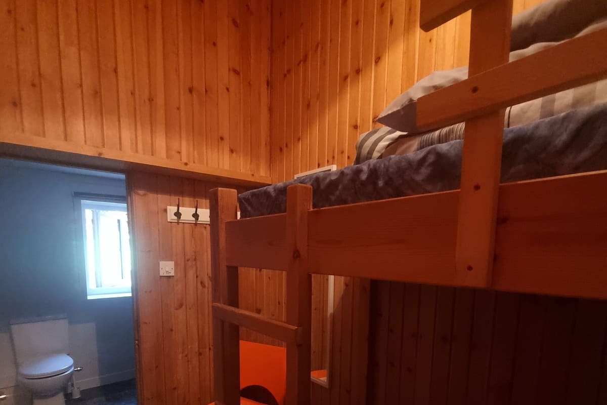 Habitación con literas de madera, una ventana y un inodoro visible al fondo.