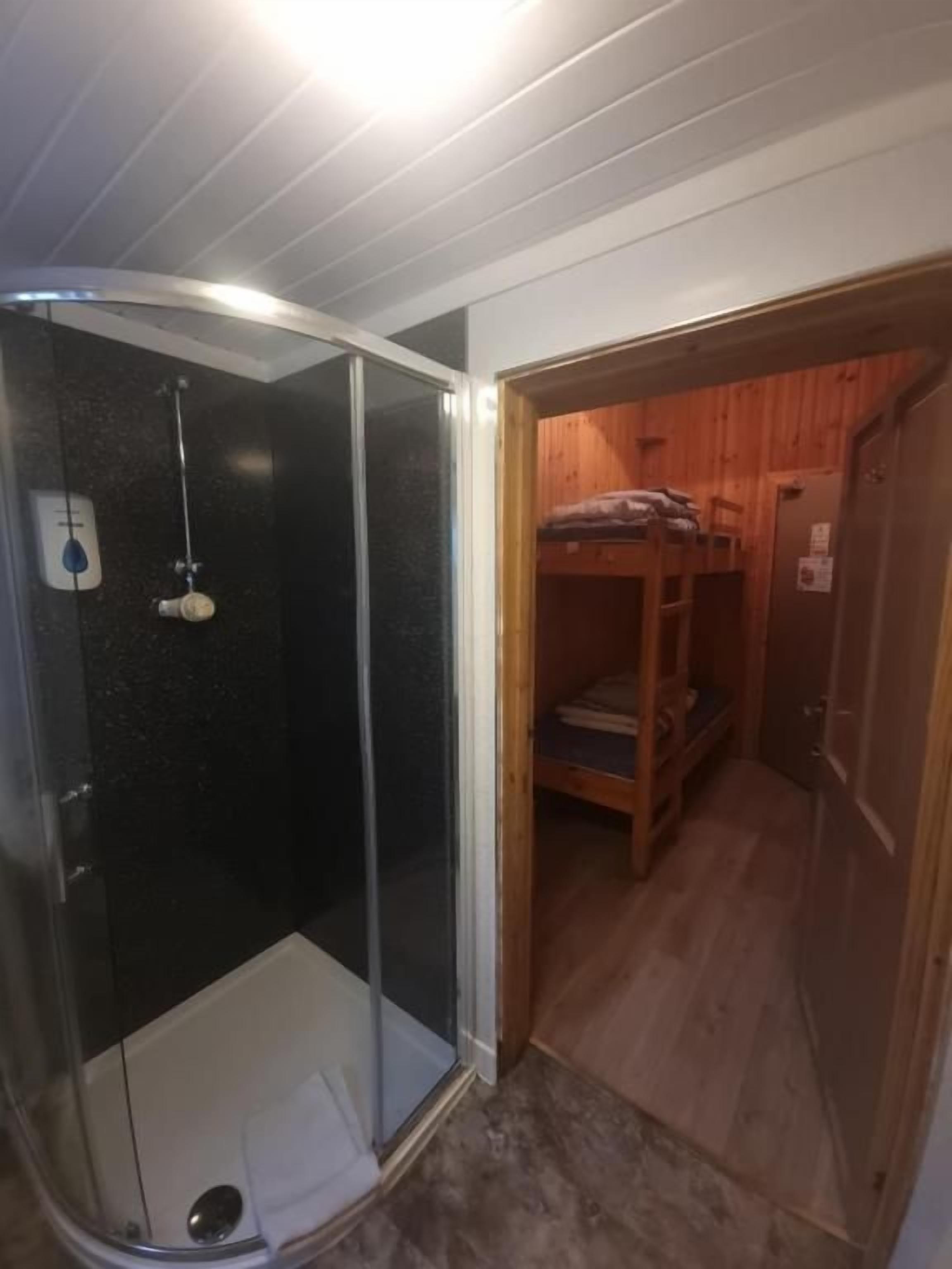 Aseo con cabina de ducha de vidrio, puerta de madera que da a un cuarto con literas y un inodoro a la vista.