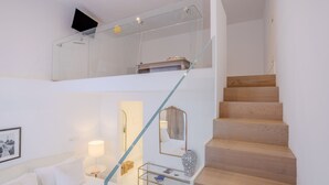 Una habitación de hotel moderna con una escalera de vidrio, una cama con ropa blanca, una mesita de noche con una lámpara y un televisor montado en la pared.