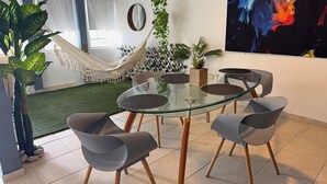 Un salon moderne avec une table en verre, quatre chaises grises, un hamac et diverses plantes.