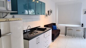 A modern kitchen with white cabinets, a black countertop, and teal upper cabinets. There is a white refrigerator, a stove, and a sink. A brown sofa and two white chairs are against the wall. - TrevizZo N.urban Pinheiros (São Paulo)
