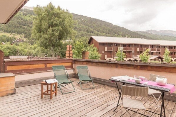 Una terraza de madera con asientos al aire libre, con vistas a un paisaje montañoso y un edificio con varios balcones.