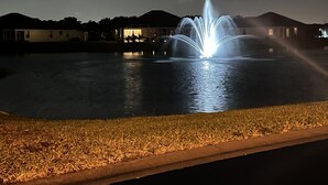 A nighttime view of a fountain with water jets, surrounded by a body of water and a grassy area with a paved walkway. - Lakeview Lanai in Fort Myers! Gated community with resort like amenities!!! (Fort Myers)